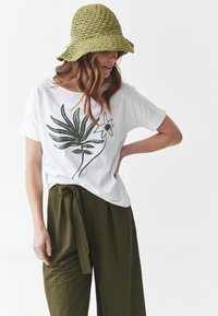 White t-shirt with green plant and flower graphic, paired with olive green, wide-legged pants. Accessorized with a textured, green woven hat.
