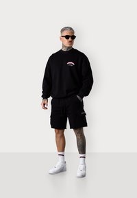 Sweatshirt noir avec un logo graphique rose ; shorts cargo noirs ; baskets blanches ; lunettes de soleil noires ; tatouage sur la jambe droite ; fond uni.