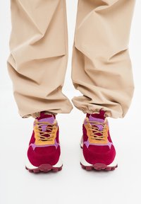 Sneaker colorati con suede rosso, dettagli arancioni e viola, mesh testurizzato e una robusta suola bianca. I pantaloni sono beige con polsini elasticizzati.