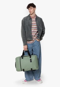 Borsa in tessuto verde con accenti neri, forma rettangolare, manici superiori e chiusura con cerniera. Il modello indossa una camicia a righe e jeans a gamba ampia.