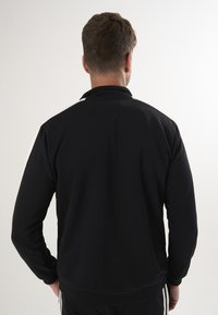 Schwarze Pulloverjacke mit Kragen, langen Ärmeln und enganliegendem Design. Mit weißen Streifen entlang des Kragens und der Seitennähte.