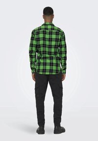 Grünes und schwarzes karierter Flanellhemd mit langen Ärmeln, Knopfmanschetten und geradem Saum, getragen mit schwarzen Cargo-Hosen und Stiefeln aus der Rückansicht.