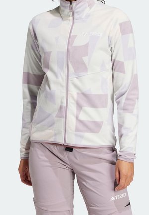 Femme portant une veste polaire zippée à motif violet clair et blanc, avec un pantalon Terrex violet assorti, debout les mains détendues.