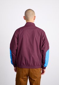 Burgundy bombarjacka med blå detaljer på ärmarna, elastiska cuffs och nederkant. Slät textur, lättviktsmaterial, inga synliga mönster eller hårdvara.