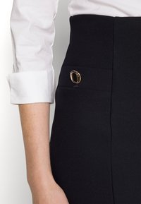 Jupe noire taille haute en tissu texturé, dotée d'un détail en bouton doré sur le côté et d'une finition lisse.
