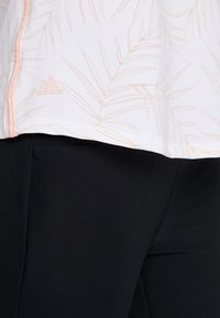 Närbild av en vit sportjacka med ljusrosa palmbladsmönster och Adidas-logga, kombinerad med svarta träningsbyxor.