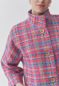 Veste tissée texturée en rose et bleu, avec un col haut, trois boutons décoratifs et un motif à carreaux.