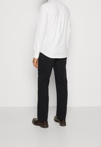 Witte longsleeve-shirt met knoopmanchetten, met een afgeronde zoom. Gepaarde met donkere broek en bruine schoenen, gezien vanuit de achterkant.