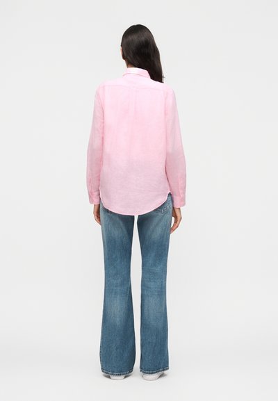 Femme debout, vue de dos, portant une chemise à manches longues rose clair et un jean évasé bleu avec des chaussures blanches sur un fond uni.