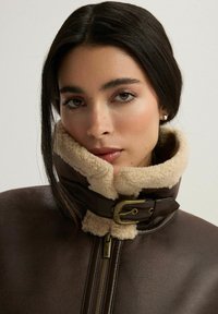 Brązowa skórzana kurtka z wysokim, beżowym sztucznym kołnierzem z owczej skóry, z detalami w postaci złotego zapięcia i zamka błyskawicznego. Gładka tekstura, dopasowany krój.