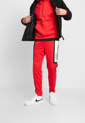 Rote Kapuzenjacke mit einer Fronttasche, kombiniert mit roten Jogginghosen, die schwarze und weiße Streifen an den Seiten haben. Darüber ist eine schwarze, strukturierte Jacke geschichtet.