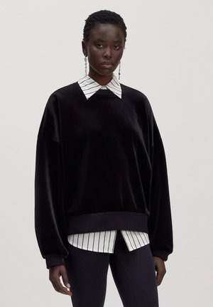 Sweat-shirt oversize en velours noir porté sur une chemise blanche avec col et rayures noires verticales sur une personne debout.