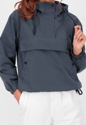 Kobieta nosząca granatową kurtkę anorak z troczkami i kieszeniami na zatrzaski, w połączeniu z białymi spodniami, ręka trzyma sznurek kurtki.