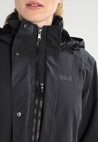 Femme portant une veste noire Jack Wolfskin avec double fermeture éclair et rabat coupe-vent boutonné, montrant le haut de la poitrine et la zone de la bouche.