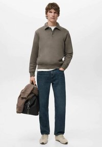 Dunkelgraues Sweatshirt mit Kragen, kombiniert mit blauen Jeans und beigen Turnschuhen. Hält einen braun-schwarzen Rucksack.