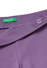 Fioletowa bawełniana tkanina z detalem guzikowym i zieloną metką z napisem "United Colors of Benetton". Teksturowana powierzchnia, widoczny detal kołnierzyka.