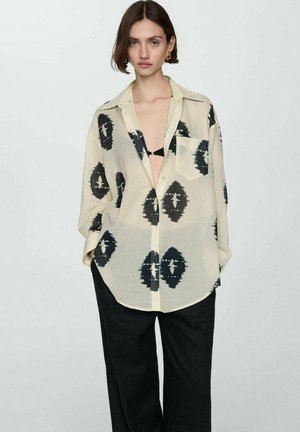 Lightweight, sheer cream shirt with black abstract patterns, collared, button-down design, and a relaxed fit. Paired with dark pants.