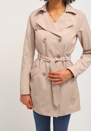 Kobieta ubrana w beżowy dwurzędowy płaszcz trencz z przewiązanym paskiem w talii, stojąca na tle jasnego, jednolitego tła.