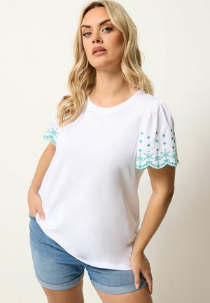 Blond vrouw die een wit blouse met korte mouwen draagt met groene geborduurde gegolfde randen en denimshorts, poserend met één hand in de zak.