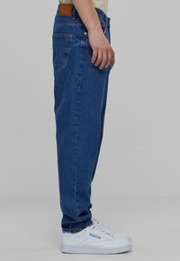 Modré džínové kalhoty s uvolněným střihem, viditelným švem a bočními kapsami. Nosené s bílými sportovními botami s zelenými akcenty.