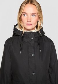 Juodas gobtuvas striukė su minkštu vidiniu pamušalu, priekiniais mygtukais ir reguliuojamomis варочkami. Lygi tekstūra su laisvu siluetu.