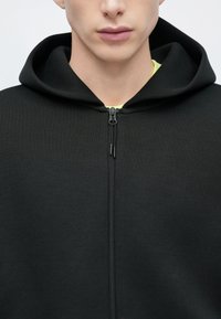 Μαύρο φούτερ με φερμουάρ και ψηλό γιακά με συνδεδεμένη κουκούλα, που διαθέτει λεία υφή υφάσματος και αντίθετη φωτεινή εσωτερική επένδυση.