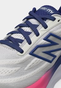Λευκό και μπλε αθλητικό παπούτσι New Balance με μπλε κορδόνια, υφασμάτινο άνω μέρος με υφή, μπλε λογότυπο "N" και λεπτομέρεια σόλας με ροζ διαβάθμιση.