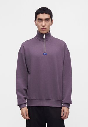 Mladý muž s krátkymi čiernymi vlasmi, ktorý nosí fialovú mikinu na zips s malým modrým logom "HUGO" na hrudi, stojaci pred bielym pozadím.