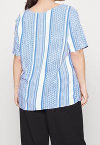 Nainen, jolla on päällään lyhythihainen sinivalkoraidallinen paita ja mustat housut, näkyy takaa yksinkertaisella taustalla.