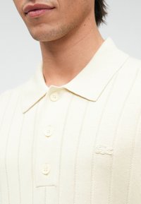 Κρεμ πόλο μπλούζα με κάθετες ρίγες, μαλακή υφή, τριπλό κλείσιμο με κουμπιά και κεντητό λογότυπο στην αριστερή πλευρά.