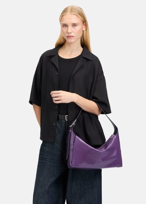 Sac à main synthétique violet, brillant, de forme courbée et avec une seule bandoulière, tenu par une personne portant une chemise noire surdimensionnée.