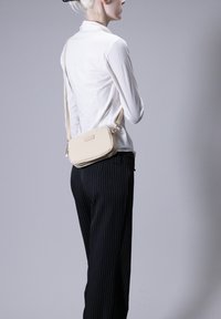 Borsa a tracolla rettangolare beige con una texture liscia, chiusura con zip e cinturino regolabile, indossata sopra una camicia bianca con bottoni e pantaloni a righe. Bottoni.