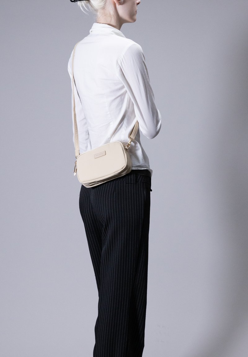 Borsa a tracolla rettangolare beige con una texture liscia, chiusura con zip e cinturino regolabile, indossata sopra una camicia bianca con bottoni e pantaloni a righe. Bottoni.