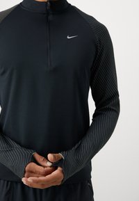 Sort langærmet træningstrøje med kvart lynlås. Har teksturerede stribede ærmer og et lille Nike-logo på brystet.