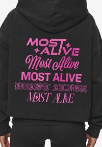 Černá mikina s kapucí s velkým růžovým textem na zadní straně: "NEJVÍCE NAŽIVU" v různých písmech a stylech, s akcenty a měkkou texturou.