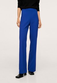 Pantalon bleu sur mesure avec une coupe droite, doté de poches latérales et d'un tissu lisse et structuré. Porté avec des chaussures noires.