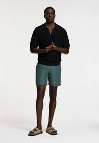 Juodas megztas polo marškinėlis, tamsiai žalsvi plaukimo šortai ir pilkos šlepetės su tamsiu padu. Modelis stovi susikabinęs rankomis, neutraliame fone.