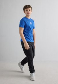 Gutt som går innendørs iført en blå Nike-skjorte med logo, svarte joggebukser og hvite joggesko mot en glatt hvit vegg som bakgrunn.