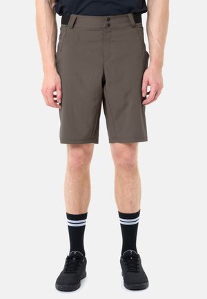 Homme portant un short mi-cuisses olive foncé, des baskets noires et des chaussettes noires montant jusqu'au mollet avec deux bandes blanches, se tenant sur un fond blanc.