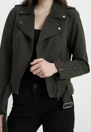 Femme portant une veste de motard vert foncé avec des fermetures éclair et des boutons argentés, associée à un pantalon noir et un haut noir.