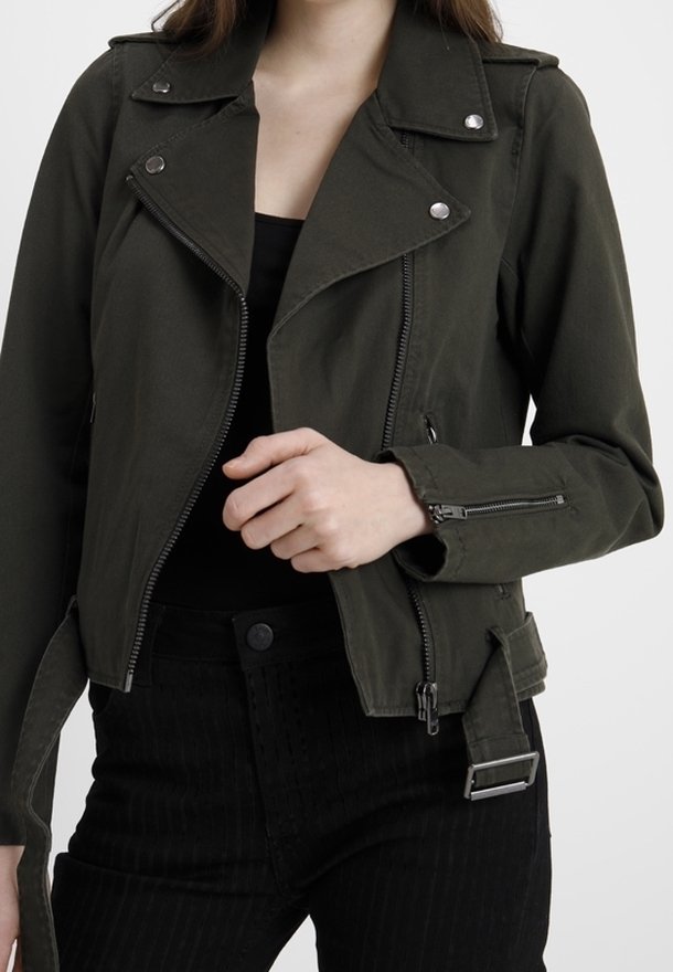 Femme portant une veste de motard vert foncé avec des fermetures éclair et des boutons argentés, associée à un pantalon noir et un haut noir.