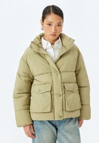 Giubbotto imbottito verde oliva chiaro con cappuccio, chiusura con bottoni, due tasche anteriori con pattina e texture trapuntata. Indossato sopra una camicia bianca.