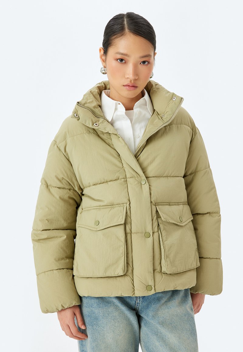 Giubbotto imbottito verde oliva chiaro con cappuccio, chiusura con bottoni, due tasche anteriori con pattina e texture trapuntata. Indossato sopra una camicia bianca.