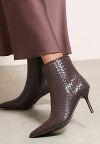 Botine negre maronii, cu o textură împletită, vârf ascuțit și tocuri subțiri. Detaliul cu fermoar pe lateral îmbunătățește funcționalitatea.