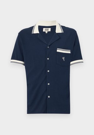 Navy sininen lyhythihainen paita, jossa on kaulus, valkoiset raidat kauluksessa ja hihoissa, teksturoitu kangas sekä rintatasku, jossa on brodeerattu kuvio.