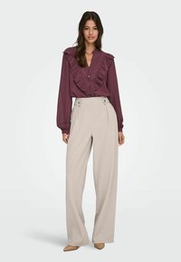 Blouse bordeaux avec un devant à volants et fermeture à boutons, associée à un pantalon large crème taille haute avec des boutons sur les côtés.