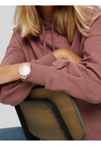 Personne en sweat à capuche mauve, reposant les bras croisés sur le dossier d'une chaise en bois, portant une montre-bracelet rose clair, avec des cheveux blonds mi-longs.