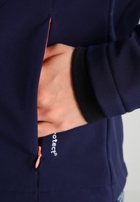 Marineblaue Softshelljacke mit Reißverschlusstasche, kontrastierendem orangen Reißverschluss und schwarzen, fleecegefütterten Bündchen; mit Logodetail am Saum.