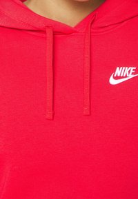Sweat à capuche Nike rouge en tissu doux avec une poche kangourou. Il comprend des cordons de serrage et un logo Nike blanc sur la partie gauche de la poitrine.