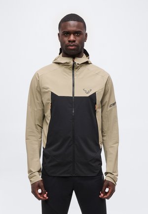 Férfi, aki bézs és fekete cipzáras kapucnis dzsekit visel, egyenes háttal áll egy egyszerű világos háttér előtt.